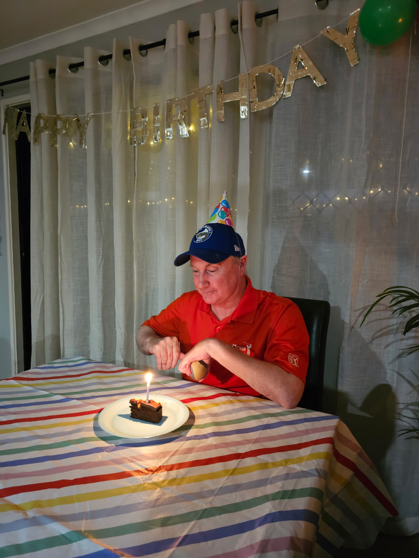A man in a party hat smiling at a slice of birthday cake with a lit candle