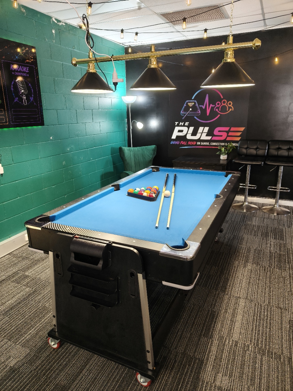 The Pulse pool table set up under the Pulse neon logo and Going Full Send mural, lit by string lights and a three-shade overhead fixture