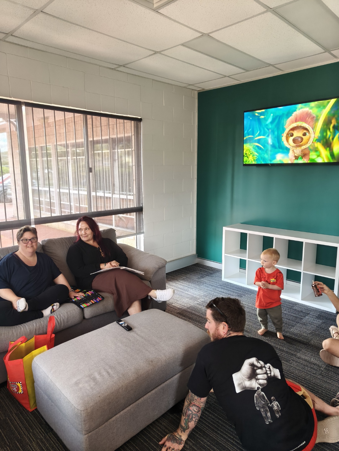 Guests relaxing together in The Pulse lounge on Open Day — some drawing at a table, a child nearby, a large TV on the teal feature wall