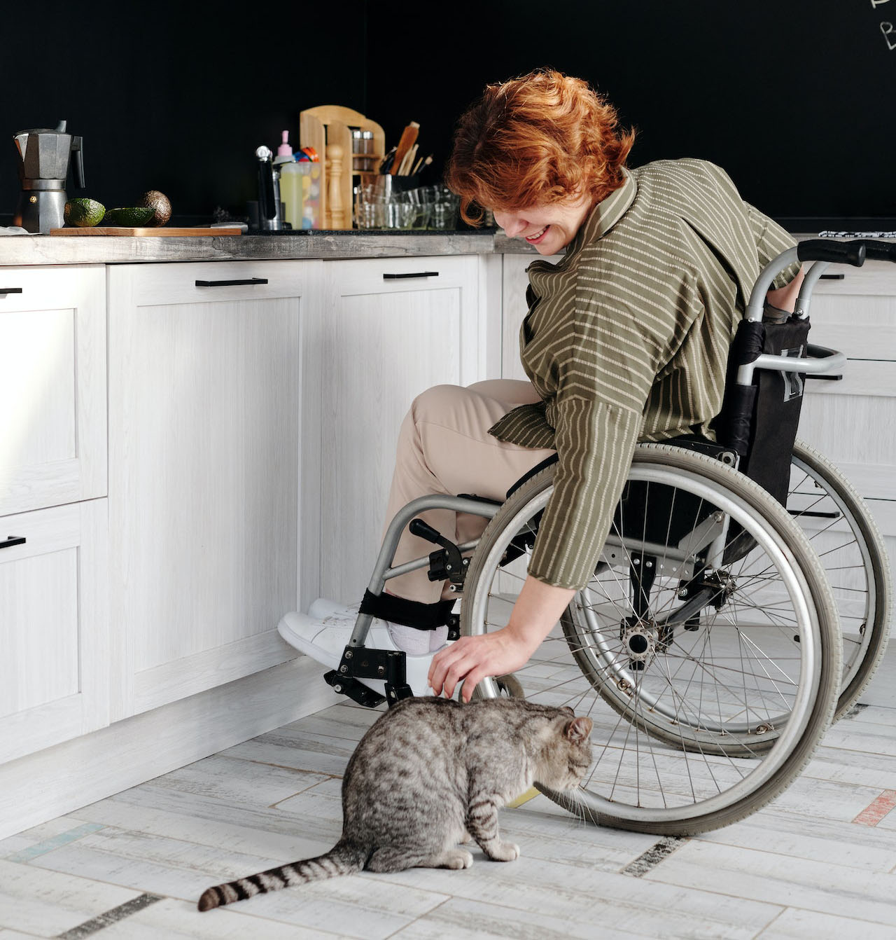 A woman sitting in a wheelchair by a kitchen bench leaning down to pet a grey cat.